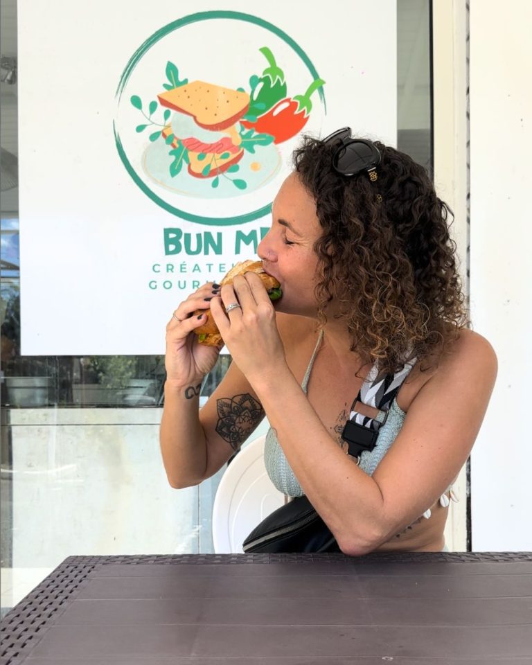 Ambre TAC dégustant un sandwich gourmand au snack Bun Me au Gosier en Guadeloupe devant le logo du restaurant spécialisé dans les sandwichs généreux et faits maison.